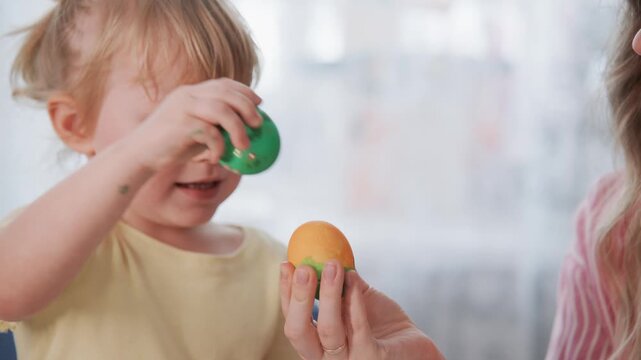 Little girl paints Easter egg with green dye. Mother holds colorful egg, smiling warmly. Child shows painted egg, eyes wide with joy. Mother watches, happy and engaged. Perfect for Easter celebrations
