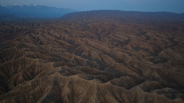 Aerial view of the rugged, arid landscape of Issyk Kul with deep ridges, undulating hills, and distant snow-capped mountains under a twilight sky in Issyk-Kul Region, Kyrgyzstan.