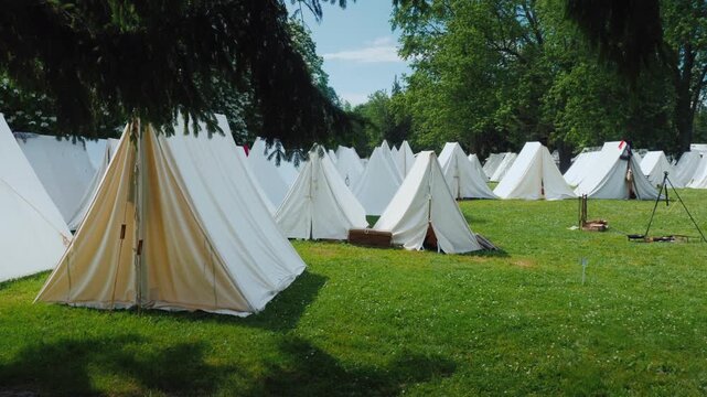 Historical white canvas tent encampment situated on grassy park field representing period military lifestyle setup outdoors within lush green trees under bright summer sun during day reenactment event