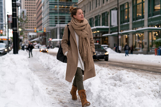 Woman strides through snowy city street. Coat, scarf, boots, and handbag complete her look. Winter's chill meets urban energy in her step. She smiles, confident, unbothered by snow
