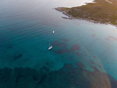 Aerial view of two sailboats floating in the clear turquoise waters of the Mediterranean Sea near the rocky coastline of Marinella, Sardegna, Italy.