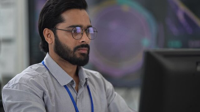 Bearded engineer with glasses and ID lanyard works at his computer under a glowing hologram display in a bright tech office, in an unposed working moment, a picture of multicultural teamwork