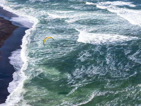Aerial view of a kitesurfer gliding over the turquoise waves near the dark sand of Depok Beach, Special Region of Yogyakarta, Indonesia.