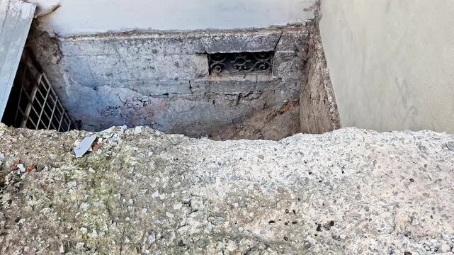 Concrete drainage channel with rough damaged walls, exposed utility opening, metal grate, and weathered surfaces, showing urban infrastructure and underground water drainage construction