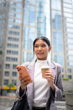 Young Asian Businesswoman Using Smartphone Holding Coffee Outdoors