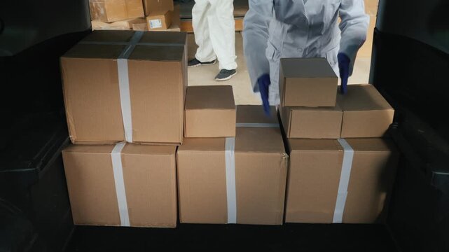 Professional worker in white uniform and protective gloves loading cardboard delivery boxes into trunk of car for transport of essential medical supplies or goods in industrial warehouse facility