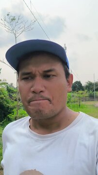 Vertical selfie-style video of an Asian man wearing a blue cap and white t-shirt walking outdoors while looking around thoughtfully, the first day jogging.