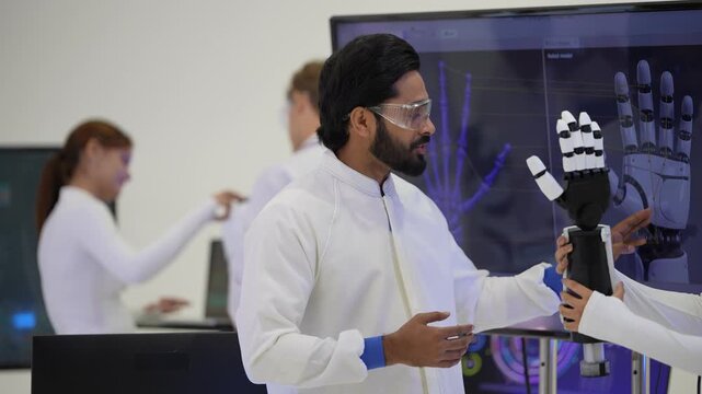 Bearded engineer in safety goggles holds a bionic robotic arm and explains it in front of a robotic hand scan display, as the team compares results, a next generation prosthetics concept