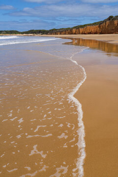 Low angled view of a foamy wave gently rolling over a golden sand beach below sea cliffs