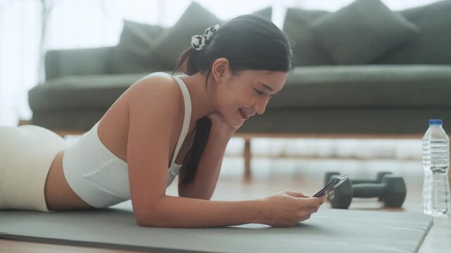 Asian woman resting after home workout and using mobile phone. Young sporty female after practicing yoga, break in doing exercise, relaxing on yoga mat, texting on the cell phone, holding smartphone