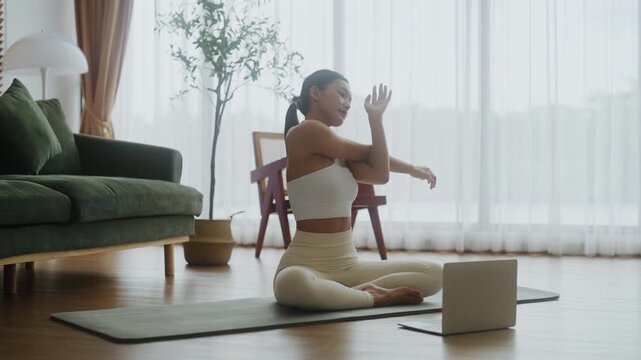 Happy young Asian woman practicing yoga and shoulder stretching at home sitting on floor in living room