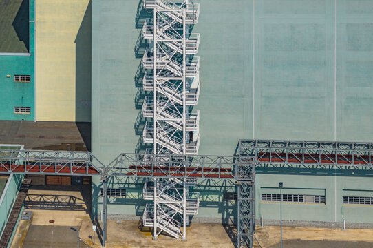Aerial view of geometric industrial architecture featuring external steel stairways, service platforms, and pipe bridges at a modern power generation facility in Chivasso, Piedmont, Italy.
