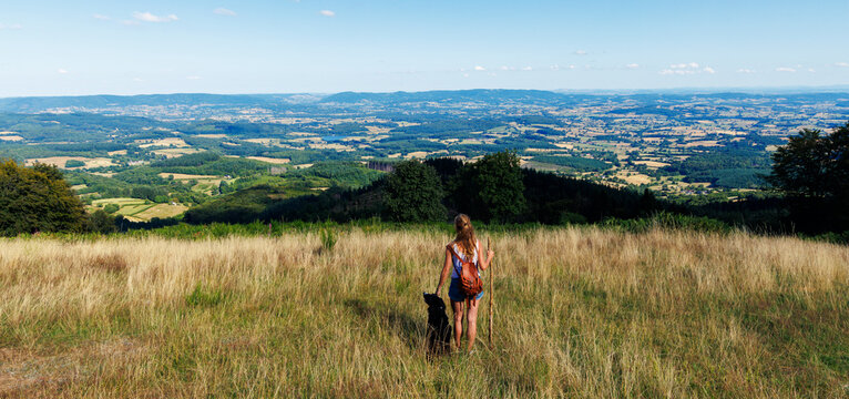 Woman hiker standing with her black dog on a hilltop looking at a vast valley landscape