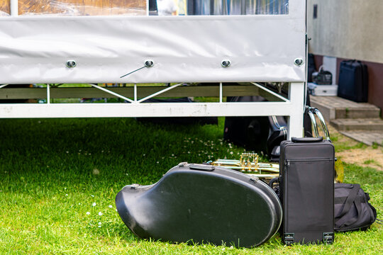 Black instrument cases and a brass French horn resting on green grass beside a white outdoor stage frame, natural daylight illumination, low angle shot.   