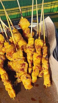 Grilled chicken satay skewers resting on brown paper inside a metal tray, filmed in a close up shot