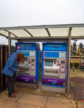  St Neots, UK - Saturday 21st October 2023: Woman using ticket machines beneath covered train station shelter beside bicycle rack during wet suburban commute with blue kiosks, touchscreen panels
