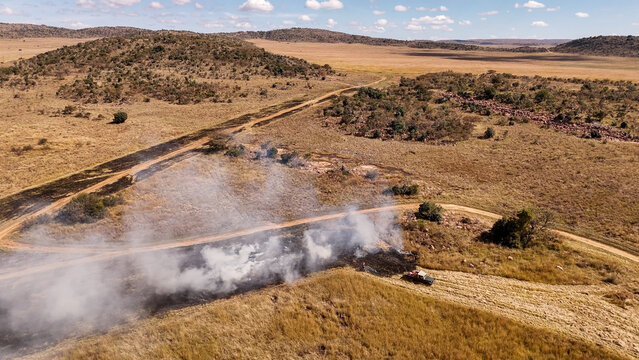 Aerial view of a controlled firebreak burn in the savanna with a vehicle nearby, dry grass, and rolling hills in the background in Modimolle, Limpopo, South Africa.