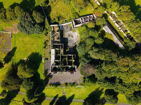 Aerial view of a derelict building with overgrown vegetation and crumbling walls, surrounded by lush green trees and a paved parking area in Lisburn, Northern Ireland, United Kingdom.