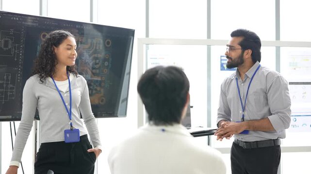 Curly-haired female engineer and bearded colleague present a circuit board design on a large screen to a coworker seen from behind, during a hands-on test, an embodied AI research scene