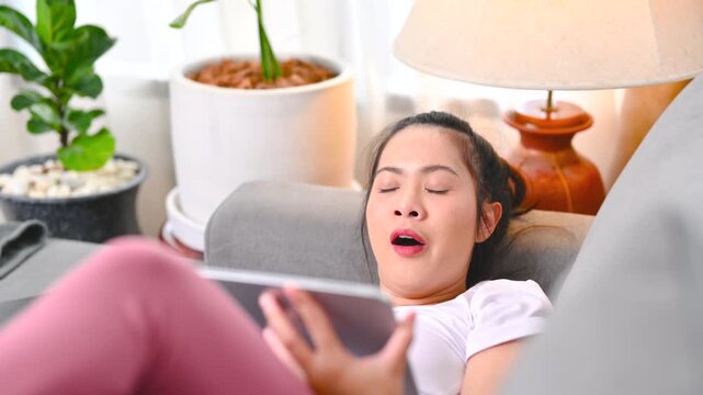 A woman holds a smartphone while wearing black headphones beside a gray sofa. The woman looks at the phone screen in the home interior; the display is too small to identify any app or service.
