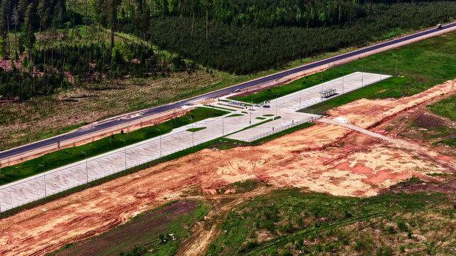 Aerial view of a paved parking area and rest stop situated alongside a road, bordered by dense forest and cleared earth in Latvia.