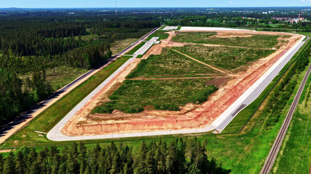 Aerial view of a large cleared land plot for industrial development, bordered by dense pine forests, a railway line, and a paved road in Latvija.