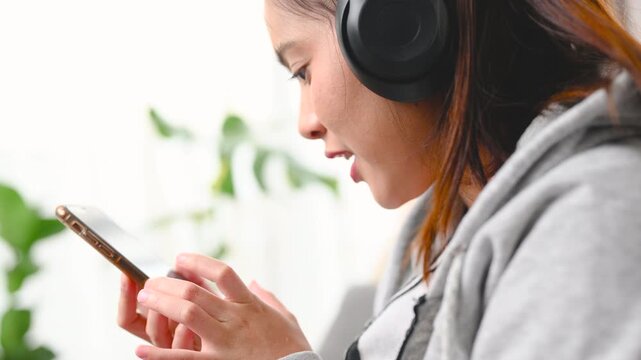 A woman holds a smartphone while wearing black headphones beside a gray sofa. The woman looks at the phone screen in the home interior; the display is too small to identify any app or service.