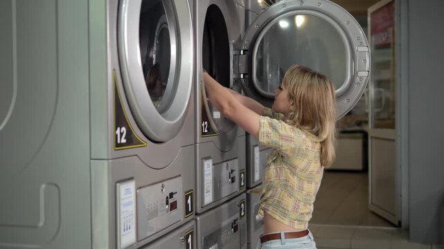 A woman loads clothes into the drum of a washing machine in the laundry room. Professional stainless steel washing machines are lined up. The process of hygienic maintenance is underway.Public laundry