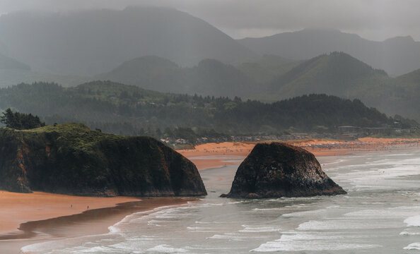 Aerial view of Haystack Rock on the sandy coastline of Cannon Beach with rolling waves, lush green coastal mountains, and a cloudy sky in Cannon Beach, Oregon, United States.