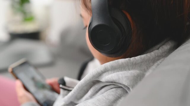 A woman holds a smartphone while wearing black headphones beside a gray sofa. The woman looks at the phone screen in the home interior; the display is too small to identify any app or service.