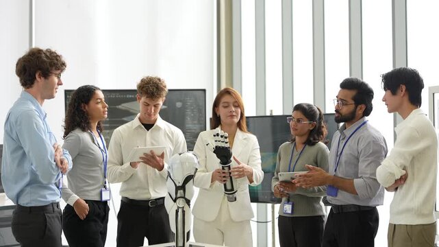 Woman in white blazer holds a bionic robotic hand as a diverse office team with tablets discusses it around a humanoid robot, during a quick check-in, a scene of real engineers at work