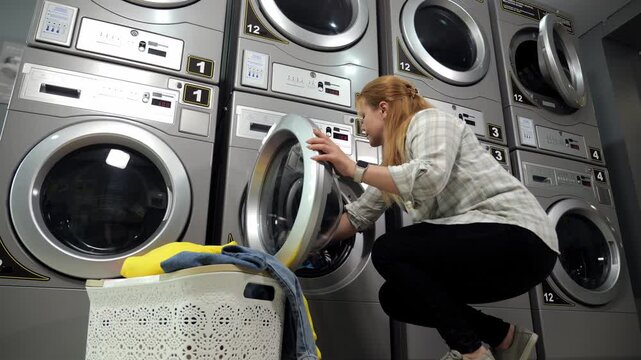 A woman loads laundry into the drum of a washing machine in the laundry room and turns on the machine by pressing the button. Professional stainless steel washing machines stand in a row.