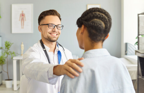 Doctor greets patient in clinic, offering support, empathy, trust, and comfort through communication and counseling. Smiling male physician gives supportive touch to African American female patient