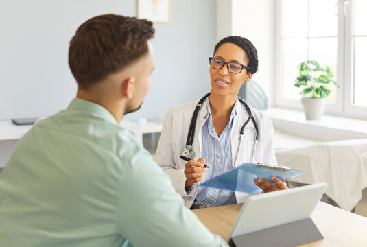 Portrait of woman physician listening and writing down patient's complaints sitting at the desk in clinic during medical examination. Sick man visiting a doctor. Healthcare and medicine concept