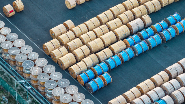 Aerial view of rows of wooden and blue-covered cable drums at an industrial storage yard, showing organized logistics and electrical infrastructure in Italy.