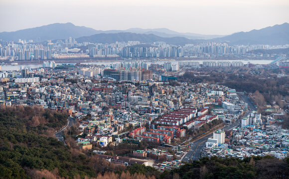 Aerial view of the dense urban landscape featuring residential buildings, high-rise apartments, and the Han River under a clear sky in Seoul, South Korea.