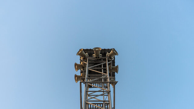 Public address sound tower and broadcast horn speaker concept, steel telecommunication tower with loudspeakers against blue sky, representing civil defense, warning alarm, and announcement.