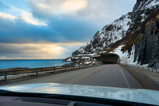 POV driving on scenic coastal highway towards an avalanche protection tunnel in Lofoten Islands, Norway. Spectacular winter road trip with snow covered mountain cliffs and ocean under dramatic cloudy 