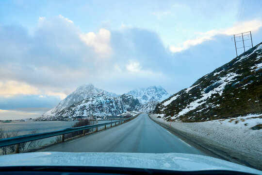 POV driving on a scenic asphalt highway during an Arctic winter road trip in Lofoten Islands, Norway. Spectacular view of snow-covered mountains and sea fjord under a dramatic cloudy sky. Beautiful Sc