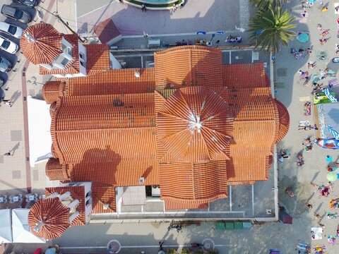 Aerial view of the Church of Agia Fotini with its distinctive orange tiled roofs situated next to a sandy beach with people and a parking area in Paralia Katerinis, Pieria, Greece.