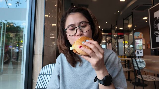 Portrait of Asian woman while eating a chicken hamburger in cafe shop. A hamburger is a sandwich consisting of a cooked meat patty on a bun or roll.