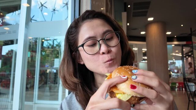 Close up of Asian woman while eating a chicken hamburger in cafe shop. A hamburger is a sandwich consisting of a cooked meat patty on a bun or roll.