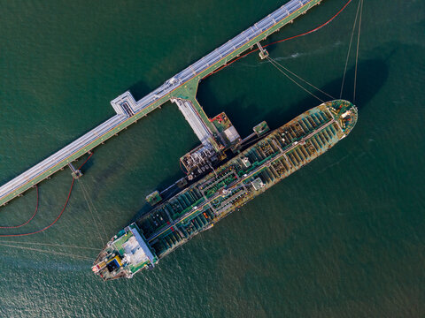 Aerial view of a large oil tanker docked at a fuel terminal pier with industrial piping and calm dark water in Cilacap Regency, Central Java, Indonesia.