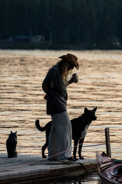 Woman in a cowboy hat drinks coffee on a dock with her dog