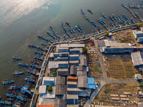 Aerial view of a coastal fishing village with numerous small wooden boats moored along the shore, featuring simple houses and drying racks in Cilacap Regency, Central Java, Indonesia.