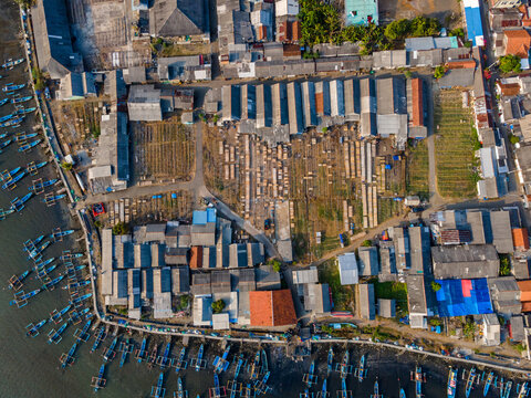 Aerial view of a coastal fishing village with small wooden boats moored along the shoreline, rows of drying racks, and dense residential housing in Cilacap Regency, Central Java, Indonesia.