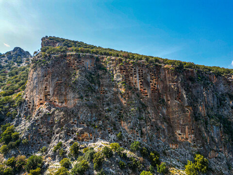 Aerial view of ancient rock-cut tombs carved into the steep cliff face surrounded by lush green vegetation under a clear blue sky in Pinararasi, Mugla, Turkiye.