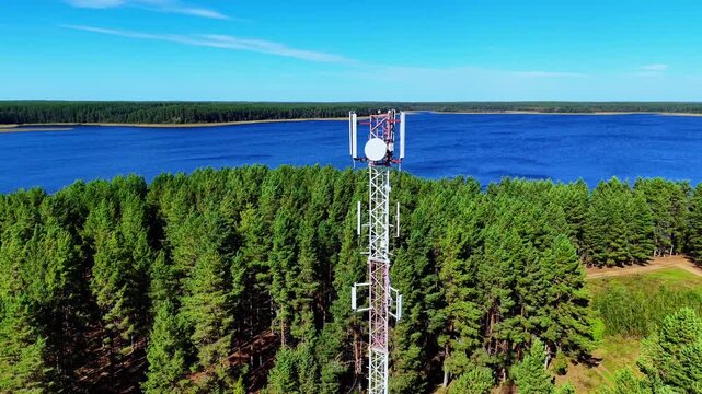 Telecommunication tower amidst lush green pine forest by calm blue lake under clear sky
