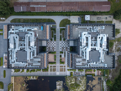 Aerial view of a modern office complex with symmetrical buildings featuring extensive rooftop HVAC ductwork, a central paved courtyard with a checkerboard pattern, and a small pond in Poland.
