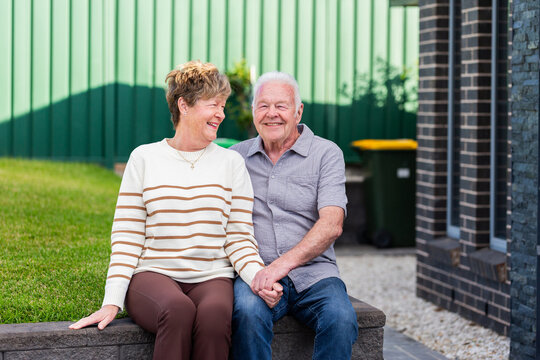 portrait of happy senior couple together sitting on retaining wall in front yard of home